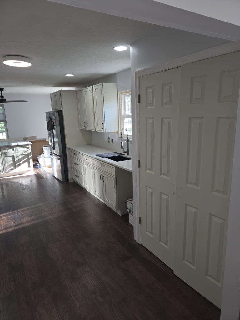 Bright, modern kitchen featuring white cabinets, a dark wooden floor, and a closed door leading to another room. Natural light streams in from a window, enhancing the spacious feel of the area.