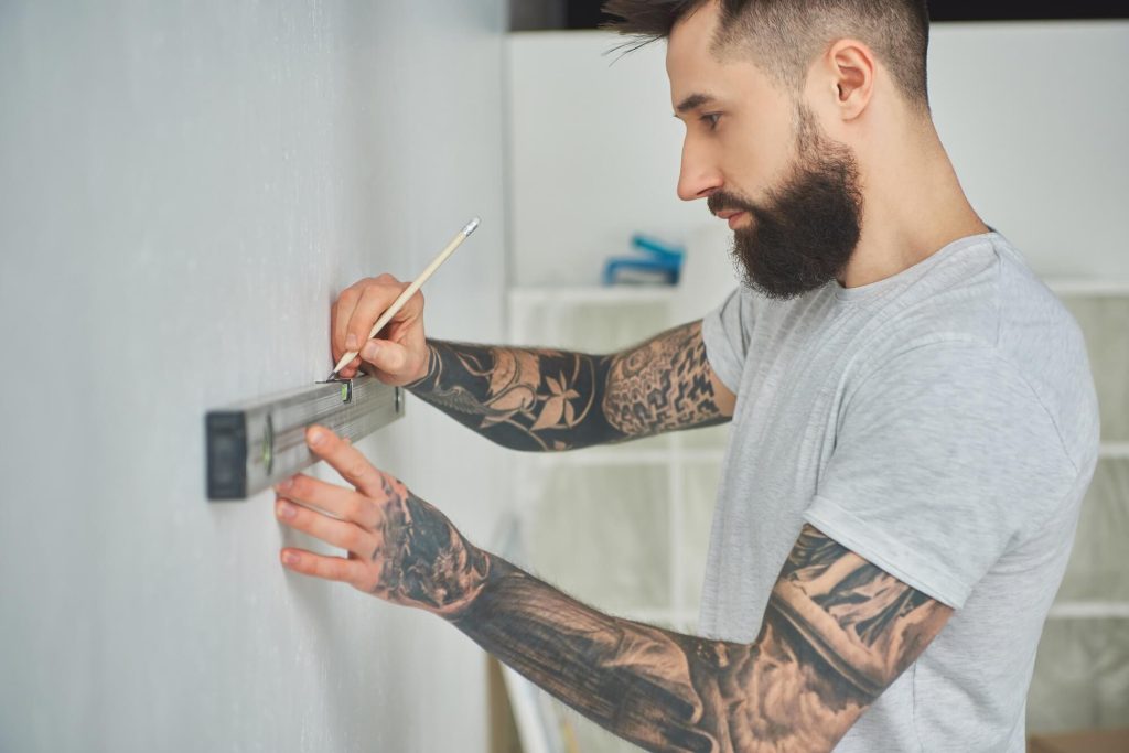 A man with tattoos is measuring a wall while preparing to paint it. He is focused on his task in a well-lit room.