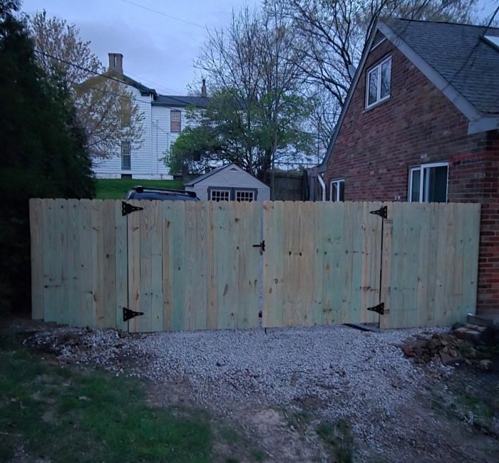 A wooden fence with a light green finish stands closed, separating a backyard from a gravel pathway. A brick house is visible to the right, with trees and another building in the background.