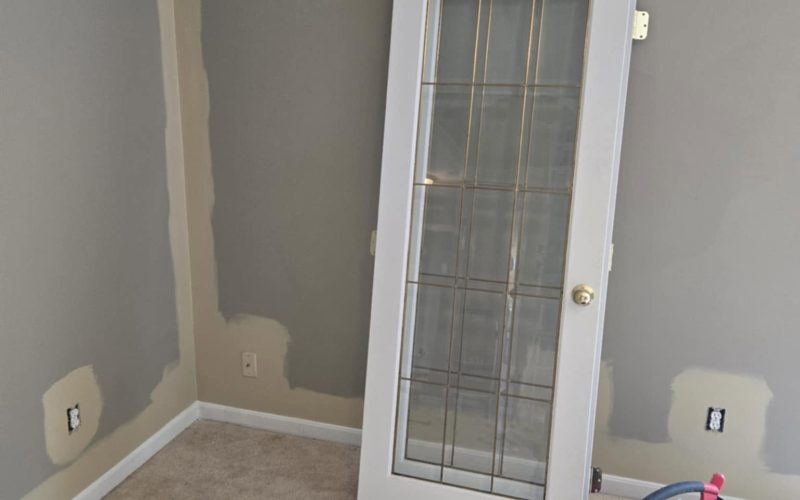 A partially painted room with a beige wall, featuring a white glass-paneled door leaning against the wall. Tools and materials are scattered on the carpeted floor.