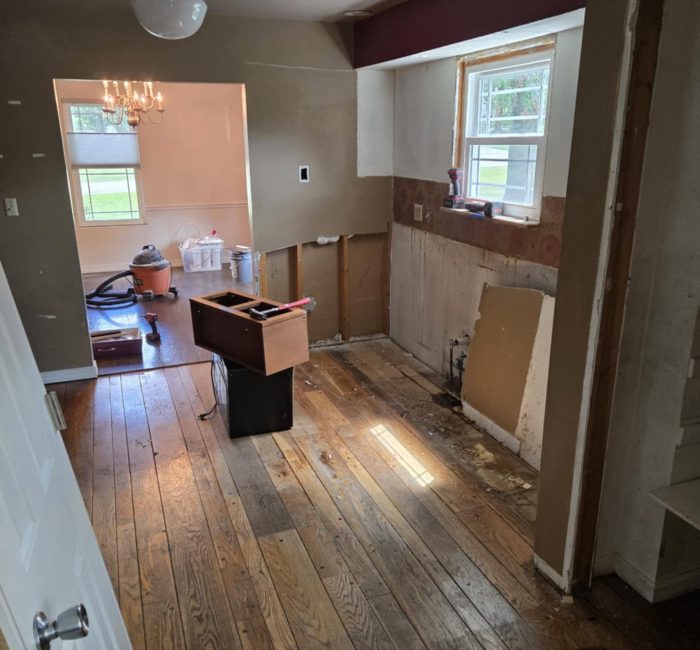 A partially renovated room with wooden flooring, showing exposed walls and a small table in the center. Natural light enters from a window, highlighting the unfinished areas.