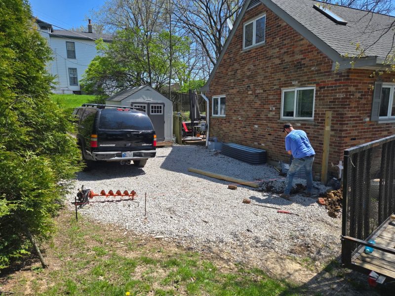 A worker is laying gravel in a yard beside a brick house, with a black SUV parked nearby. Trees and a shed are visible in the background.