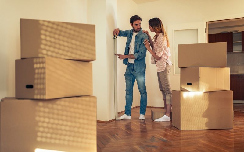 A couple discusses measurements while surrounded by moving boxes in an empty room. The scene captures the excitement of setting up a new home.